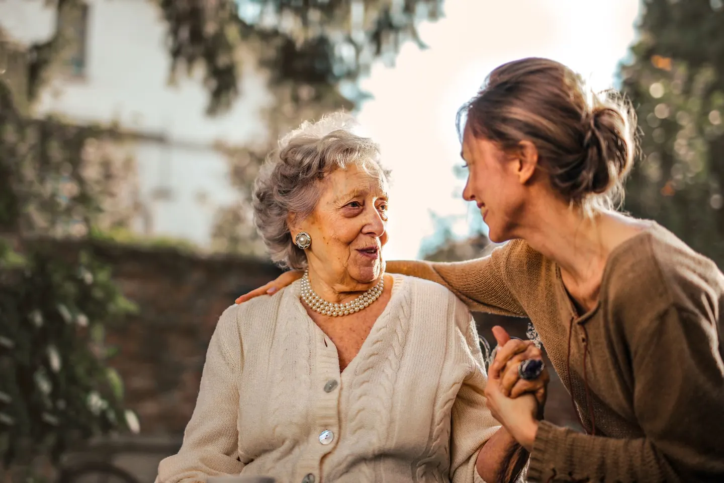 An older woman and her adult daughter sitting together in a garden, holding hands and talking.